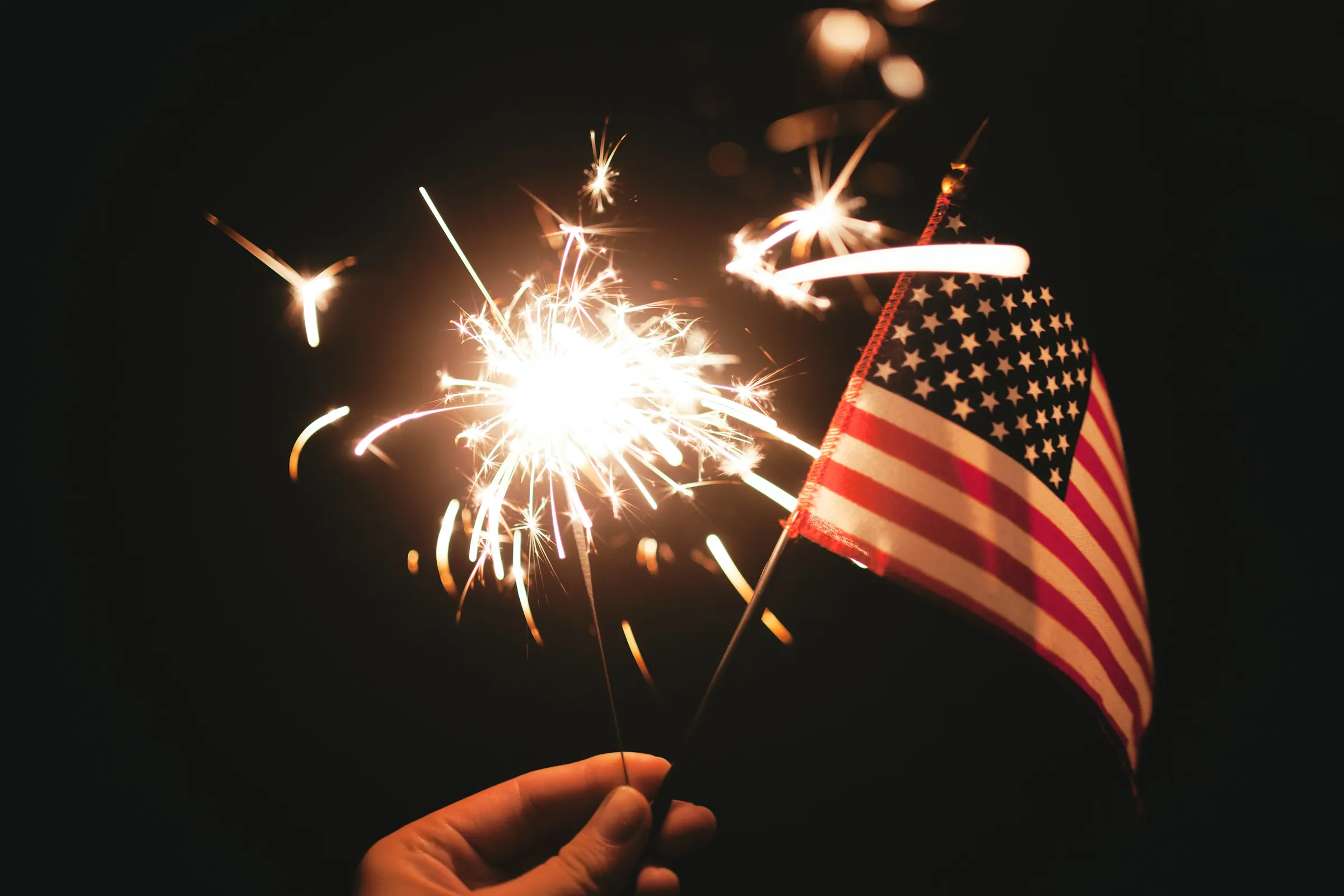 photograph of a hand holding a small American flag and a lit sparkler on a dark background