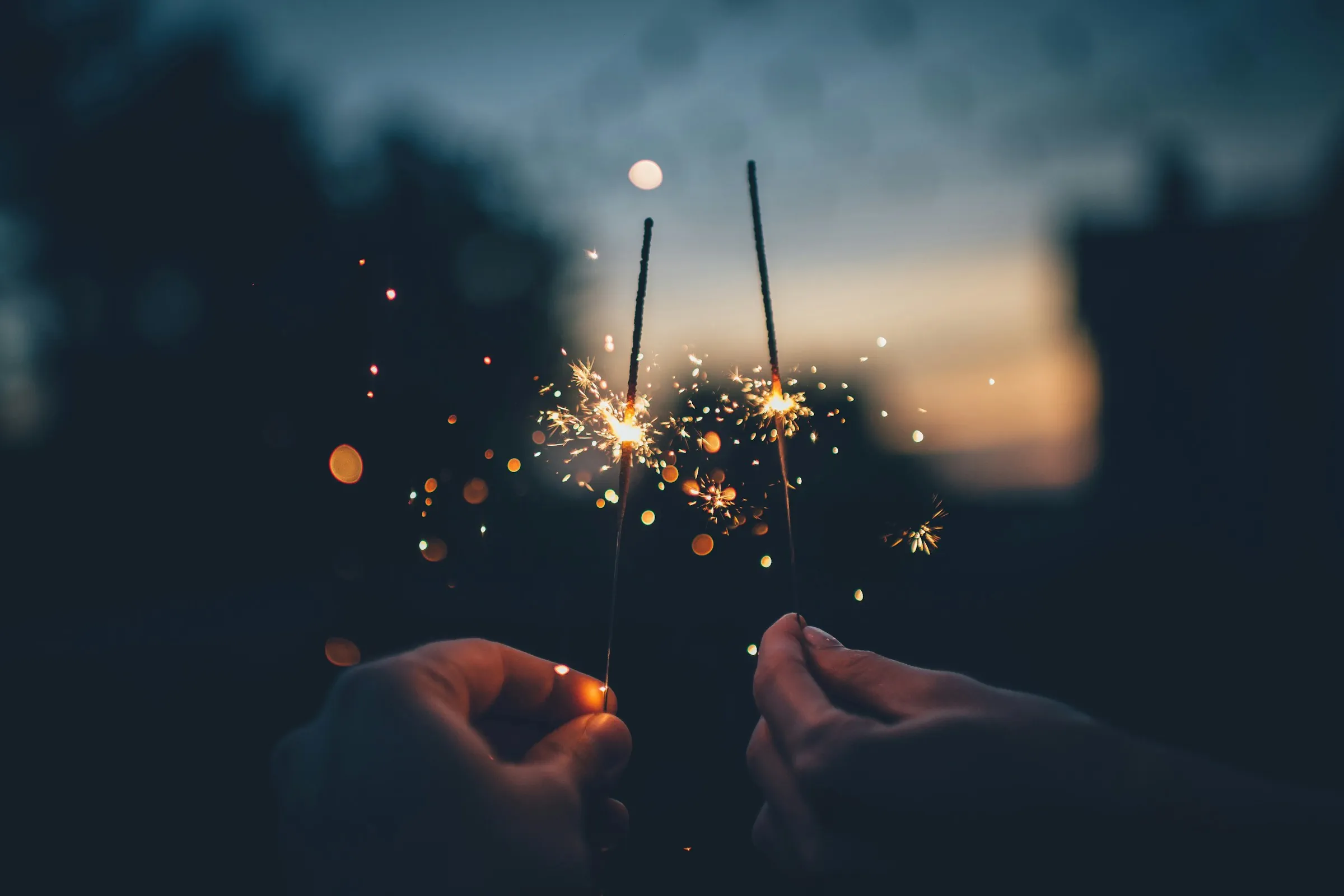 photograph of two hands holding lit sparklers on a dark background