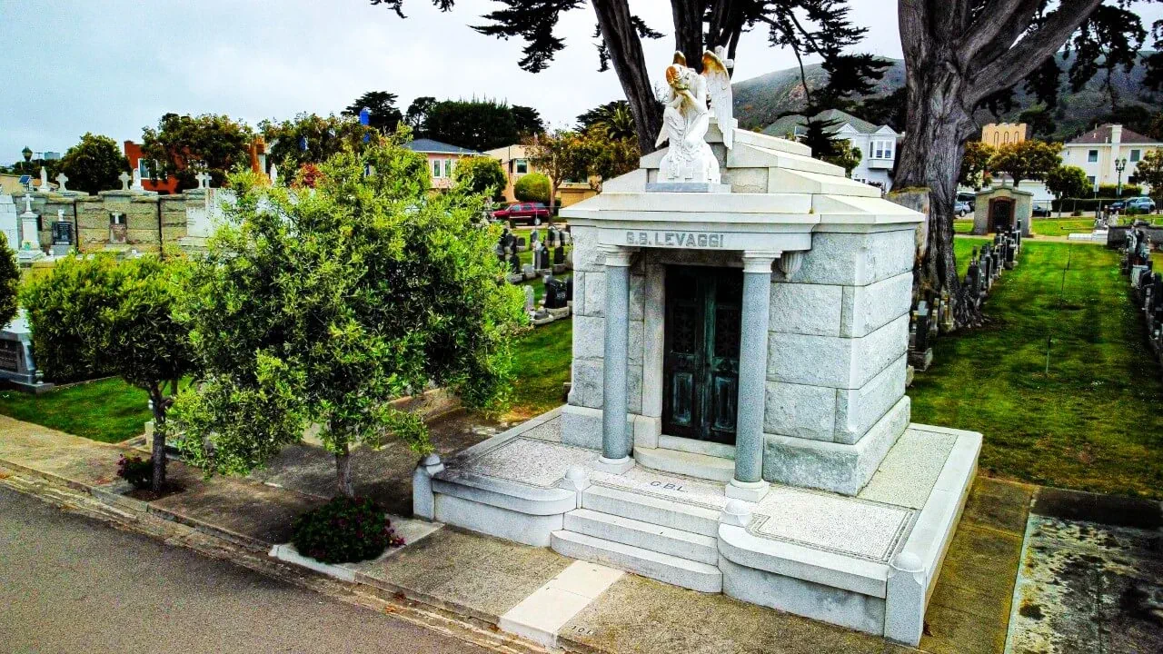 Three quarter view of a small light gray mausoleum with columns at the entrance and a large angel monument above the door