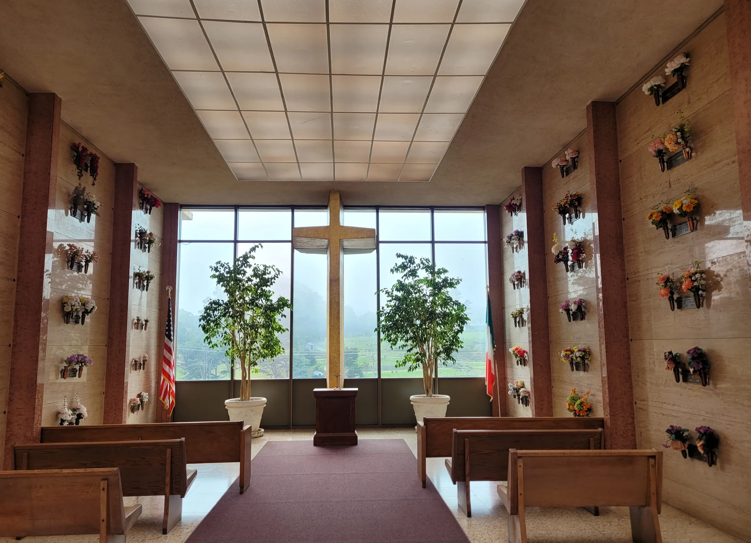 el-camino-chapel Interior view of a small private chapel featuring a large window with cross at the center flanked by potted Ficus trees