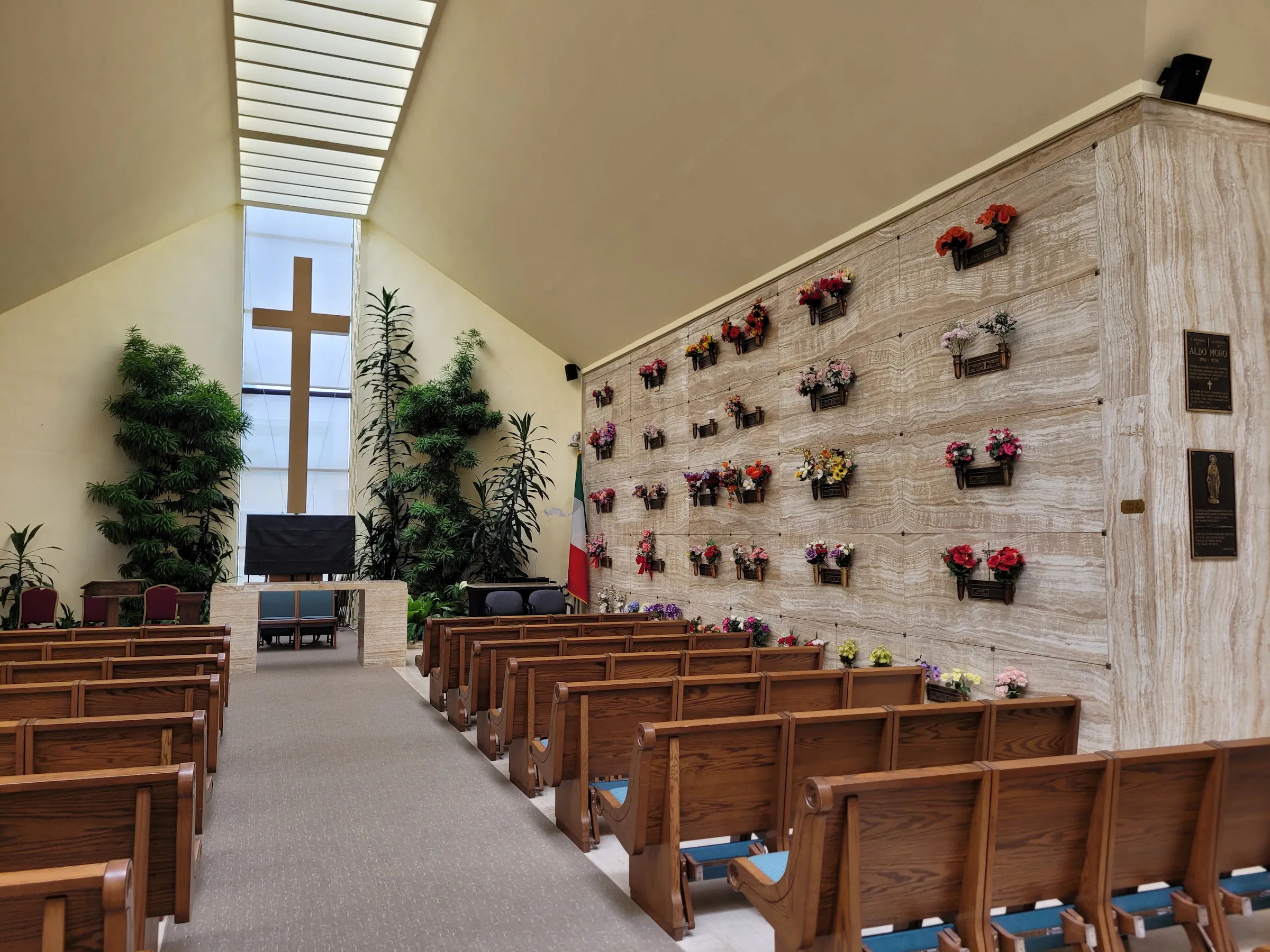 main-chapel-interior Interior view of a chapel featuring large skylights down the middle of the room, with cross at the center flanked by potted trees. Wooden pews line each side of an aisle that is centered under the row of skylights.