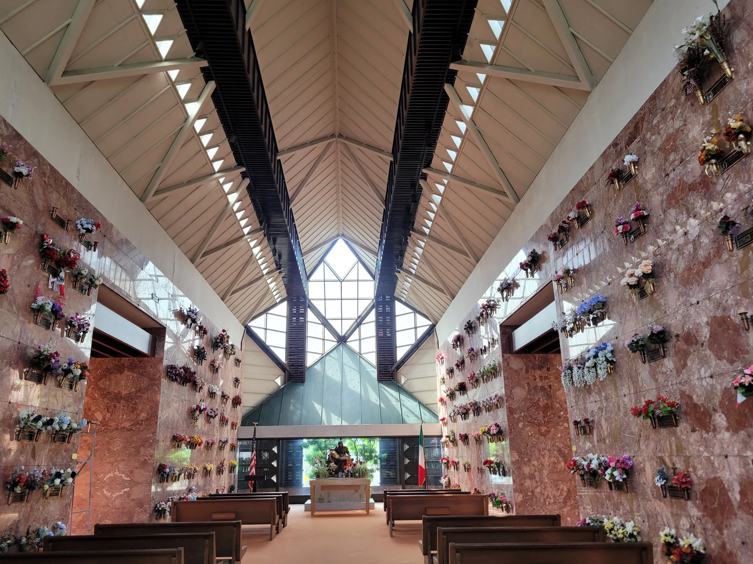 skylight-mausoleum-chapel Interior view of a chapel featuring large geometric windows. Wooden pews line each side of an aisle that is centered between walls of red granite niches.